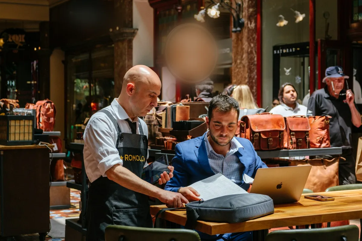 A shop owner in an apron and an adviser look through paperwork together at a laptop in a busy leather goods shop
